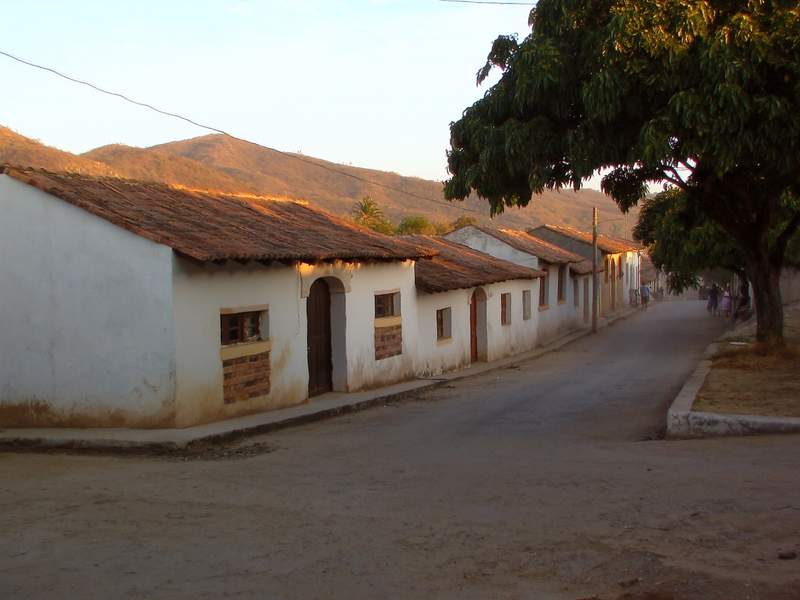 Calle ancha y polvorienta de un pueblo de calor seco, con casas bajas de adobe blanco y techo de teja y cerros pelados al fondo