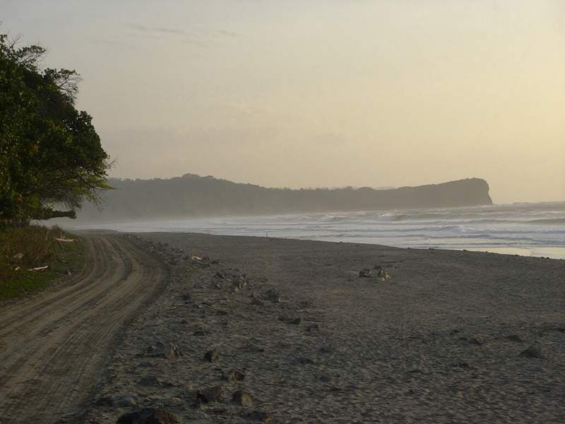 Camino de lastre paralelo a una playa larga de oleaje constante, con el monte seco de un lado y bruma marina al atardecer