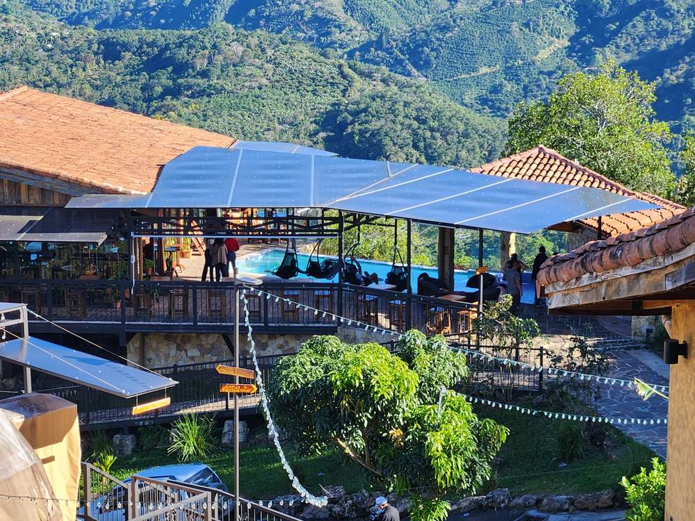 Vista amplia del restaurante y el glamping desde lejos, con la piscina, el techo de tejas y las montañas boscosas