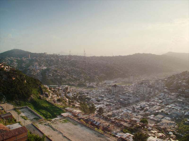 Ciudad extendida sobre un valle alto vista desde una loma, con techos bajos, quebradas verdes y la cordillera cerrando el fondo entre la calina